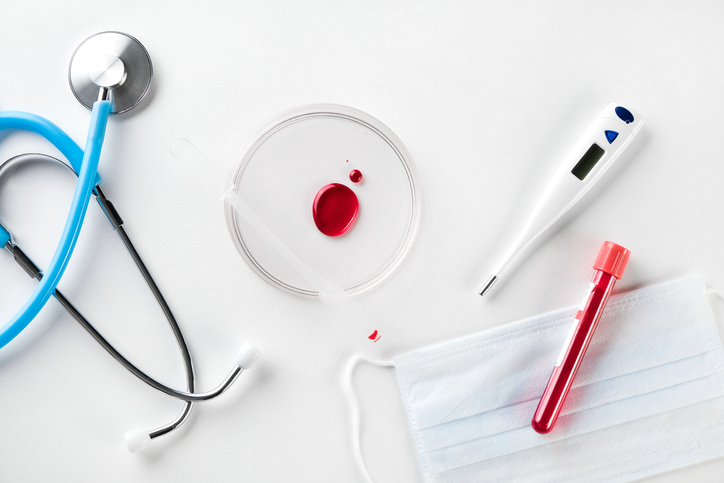 A Drop Of Blood In A Glass Petri Dish On A White Background. A Test Tube With A Medical Sample For Examination. Next To It Is A Stethoscope, A Thermometer, And A Protective Medical Disposable Mask. The concept of healthcare and medicine.
