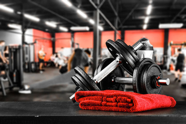 Dumbbells and red towel on dark surface, with a blurred background of a modern gym and exercising people.