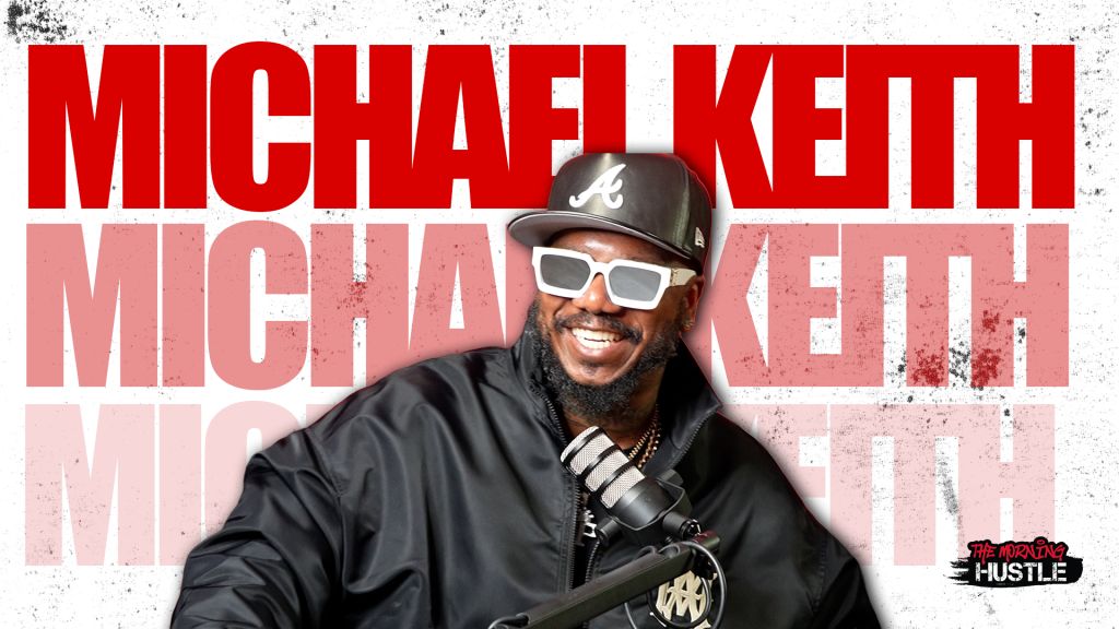 Michael Keith, a smiling man wearing a baseball cap and sunglasses, holding a microphone.