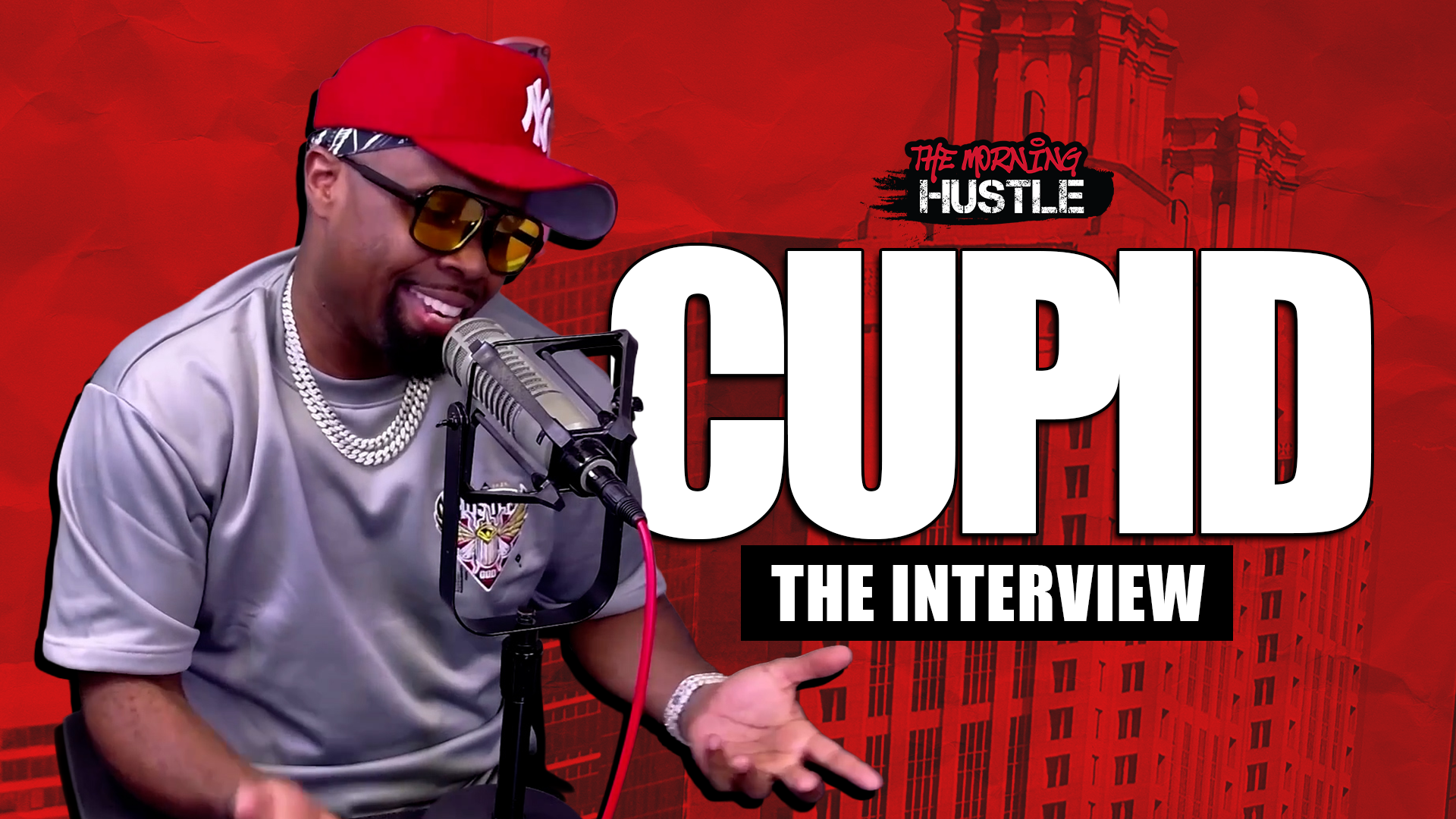 A smiling man wearing a red Yankees cap and sunglasses, holding a microphone and speaking into it against a red backdrop with the text "CUPID THE INTERVIEW" and "THE MORNING HUSTLE".