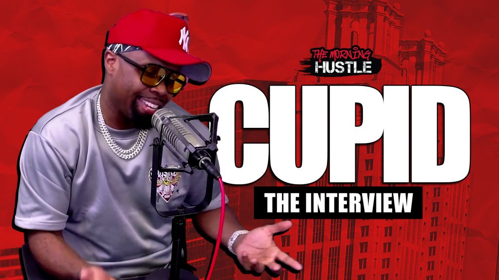 A smiling man wearing a red Yankees cap and sunglasses, holding a microphone and speaking into it against a red backdrop with the text "CUPID THE INTERVIEW" and "THE MORNING HUSTLE".