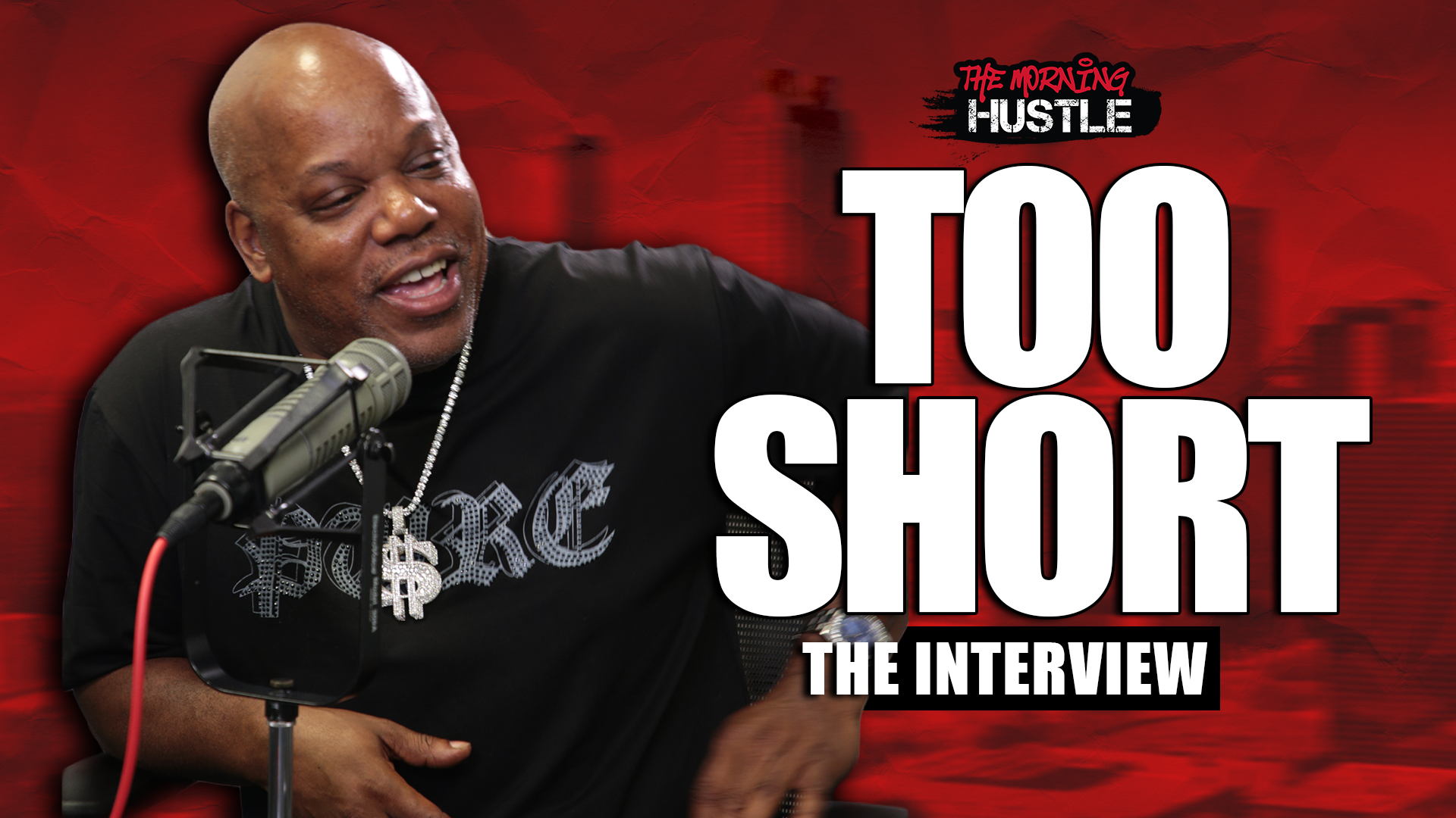 A bald Black man speaking into a microphone, wearing a black shirt with the text "The Morning Hustle" and "Too Short" printed on it.