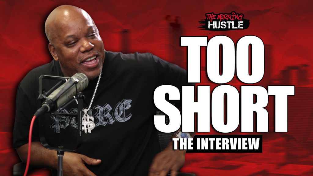 A bald Black man speaking into a microphone, wearing a black shirt with the text "The Morning Hustle" and "Too Short" printed on it.